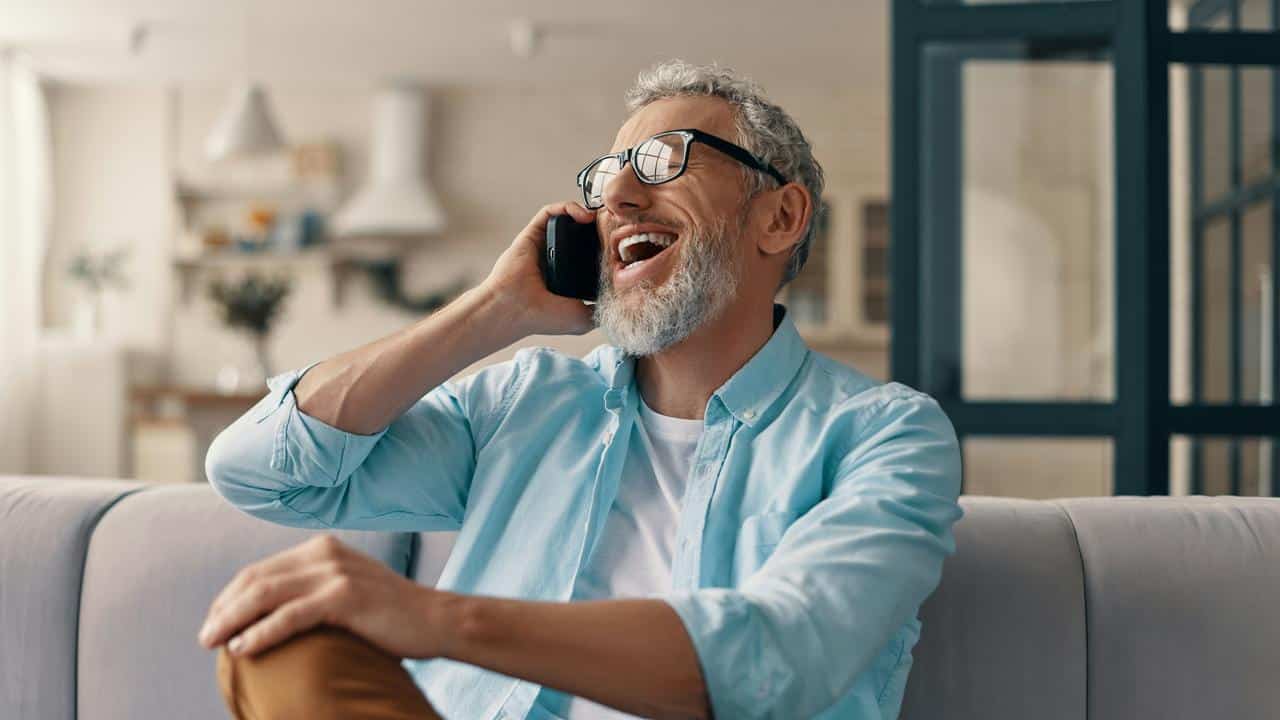 A smiling man with glasses talks on the phone at home..