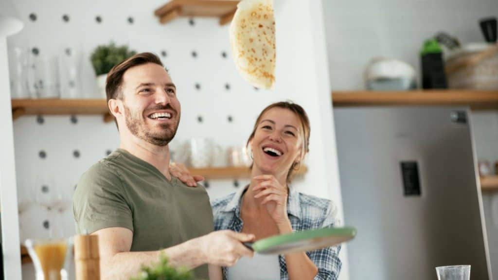 A man flips a pancake while a woman stands behind him, both laughing.