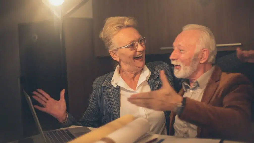 A senior couple, a woman with glasses and a man with a beard, laugh while looking at a laptop.