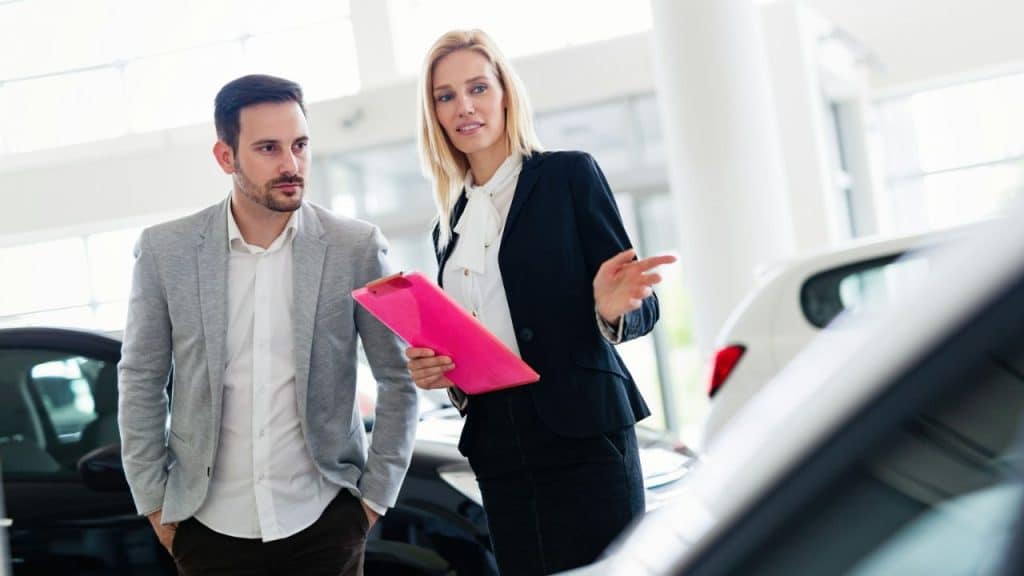 A blonde saleswoman with a pink clipboard gestures while talking to a man in a car showroom.