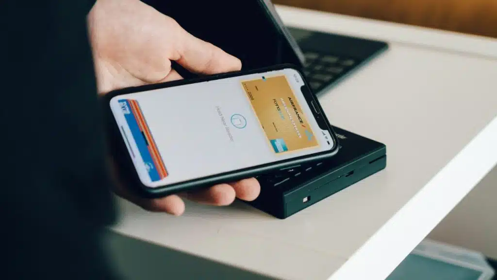 A hand holds a smartphone displaying a credit card, hovering over a black device.