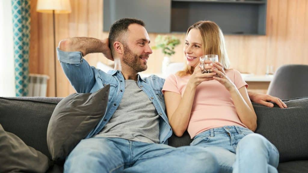 A couple smiles at each other, relaxing on a sofa with drinks