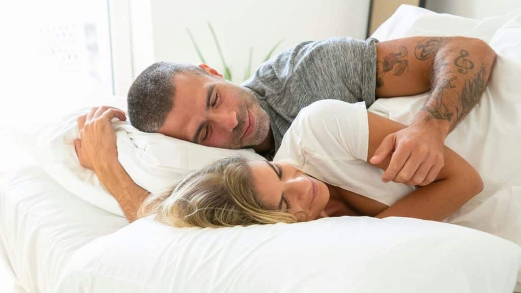 A happy couple with eyes closed, cuddling in bed under white sheets.