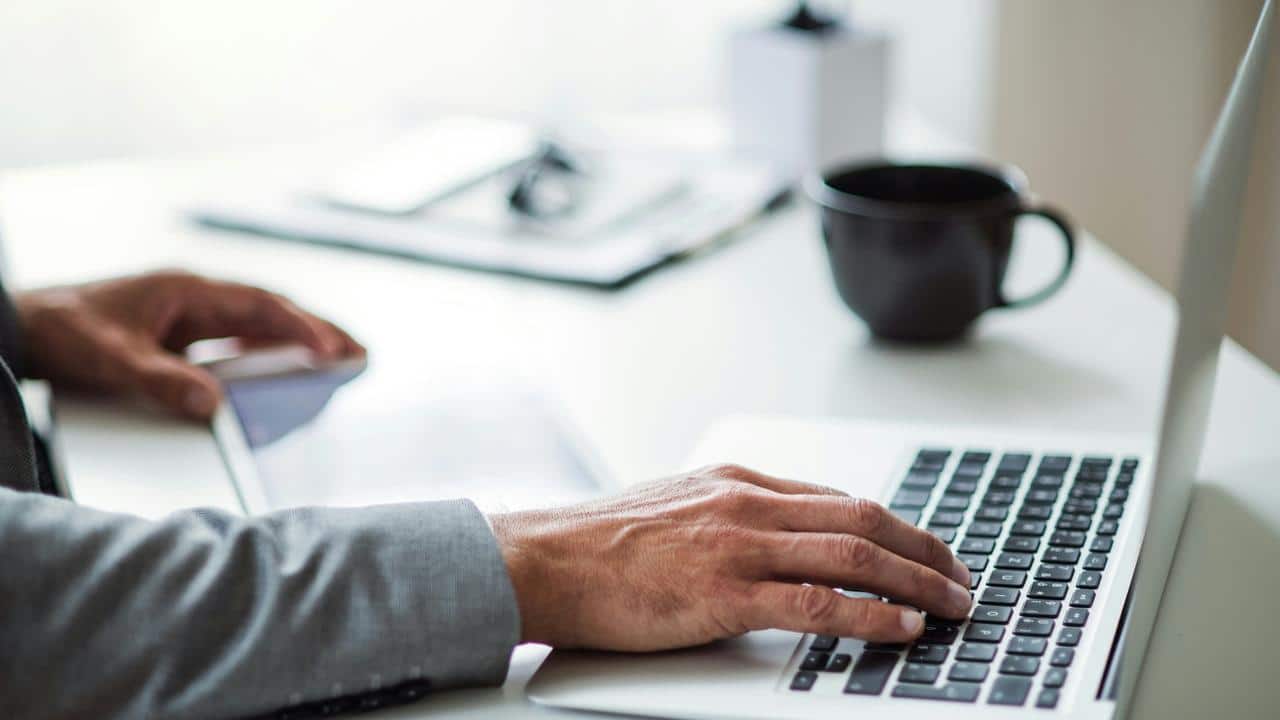 A man types on a laptop at a desk with a coffee mug.