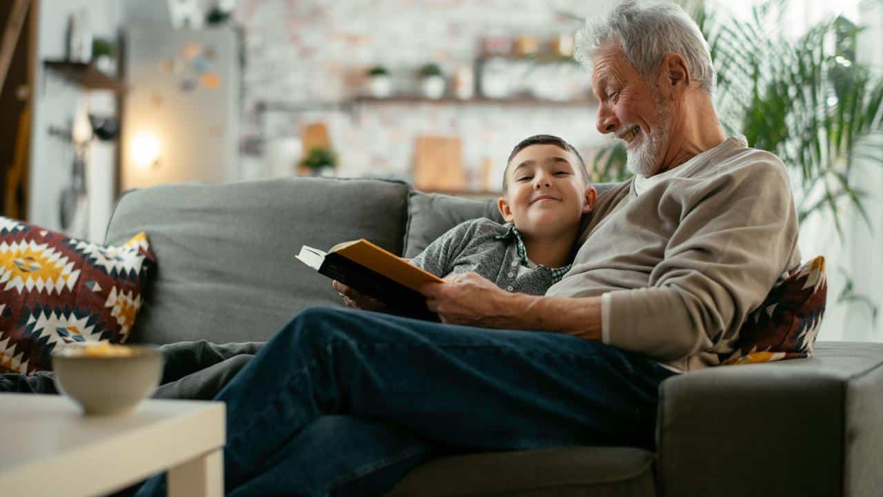 An elderly man reading with his grandson on a couch.