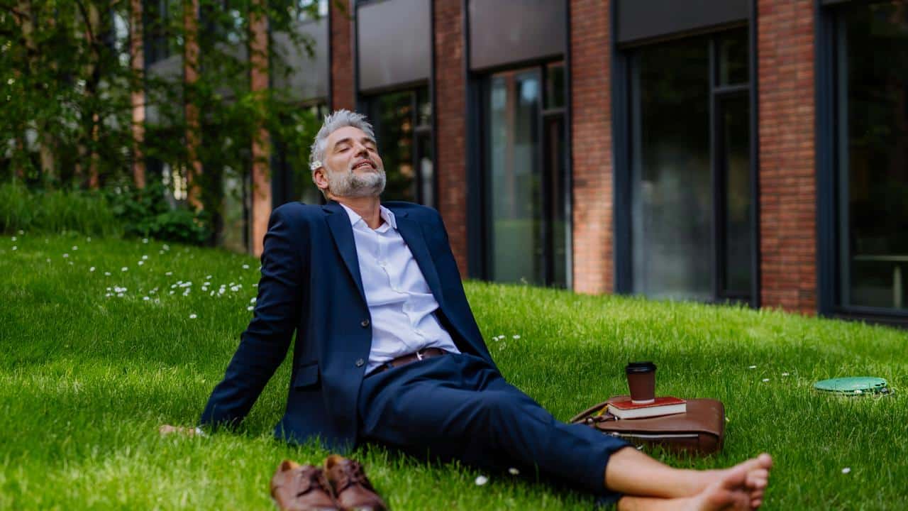 A barefoot man in a suit relaxing on grass with coffee and a briefcase.