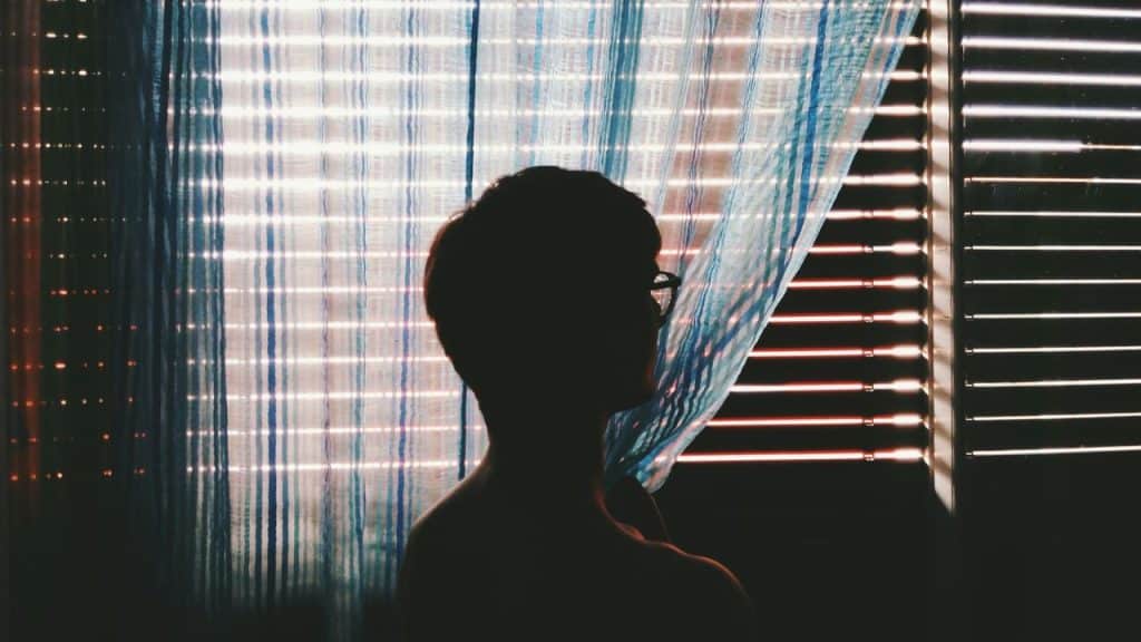 A person in silhouette with glasses looks out a window with blinds and sheer curtains.