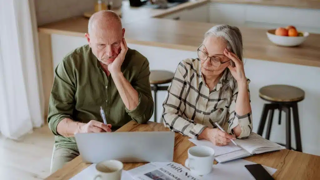 A serious senior couple sits at a table with a laptop, writing and appearing focused on finances.