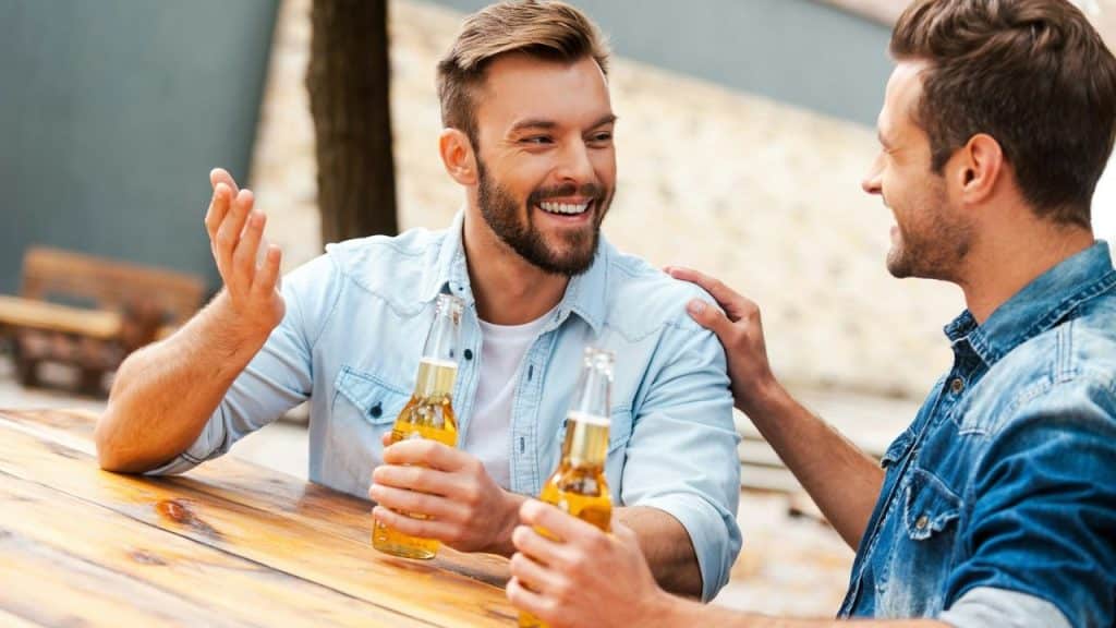A bearded man smiles and gestures while holding a beer, talking to another man with his hand on his shoulder.