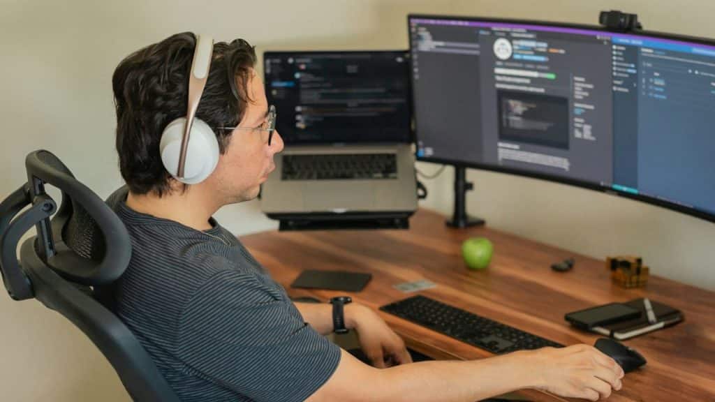 A person with headphones and glasses sits at a desk with two monitors and a laptop.