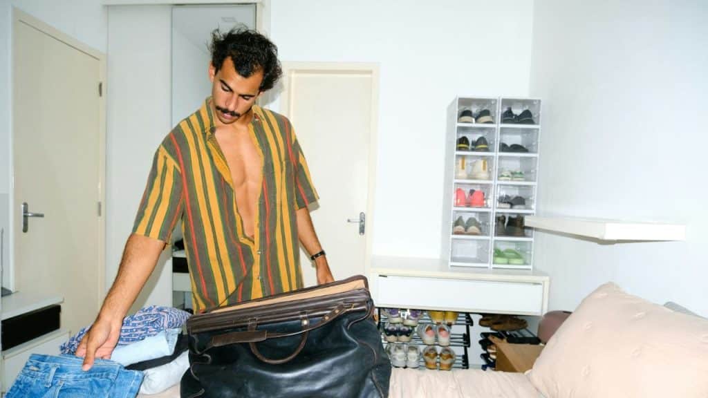 A man with an unbuttoned shirt and mustache packs a bag in a bedroom.