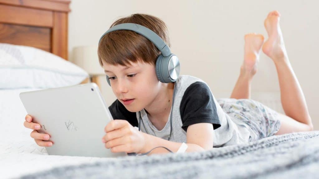 A kid wearing headphones while using a tablet on a bed.