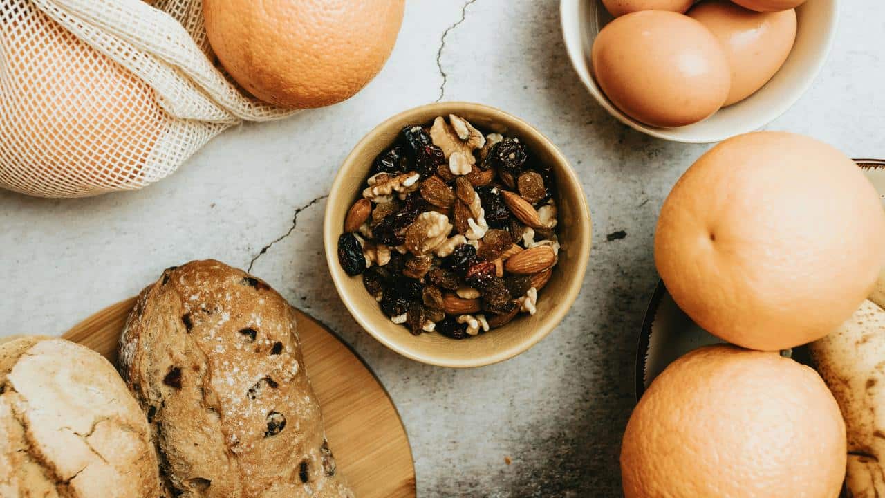 A bowl of mixed nuts and dried fruit surrounded by bread, eggs, and oranges.