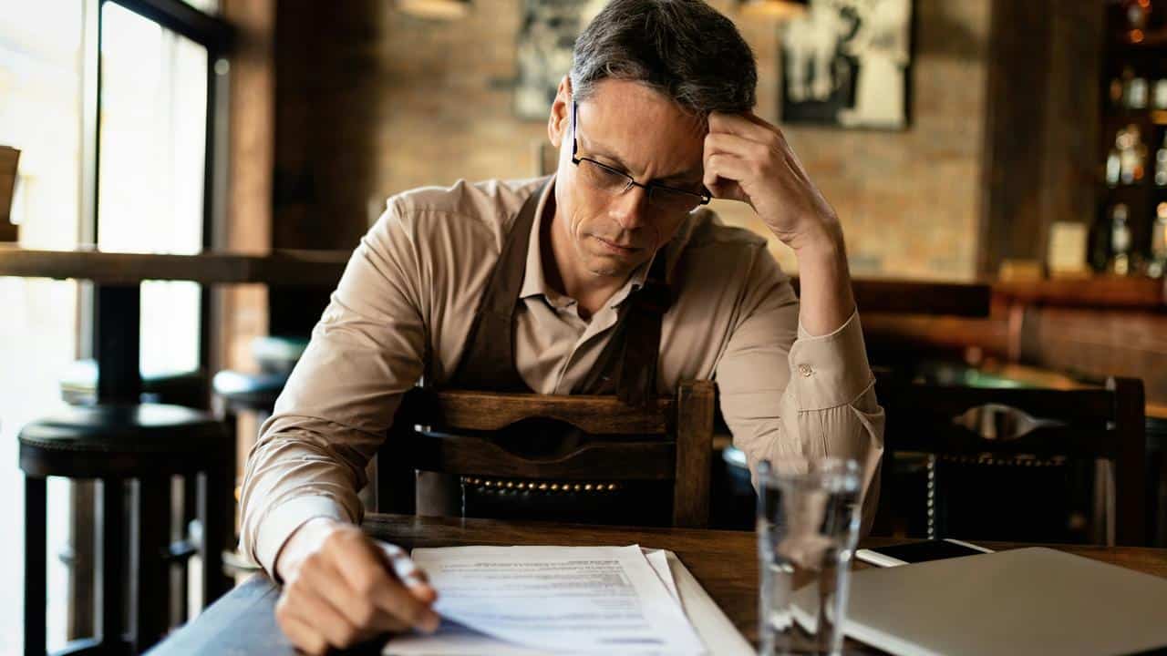 A man in an apron reviewing papers at a cafe table.