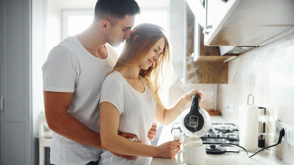 A man hugs a woman from behind as she pours from a kettle into a mug.