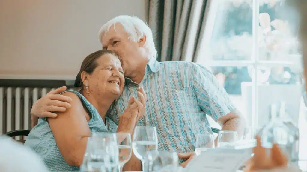 A smiling elderly couple is seated at a table, with the man kissing the woman's forehead affectionately.