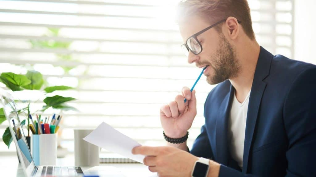 A man in a blue blazer and glasses, holding a pencil in his mouth, reviews a document at a desk with a laptop.