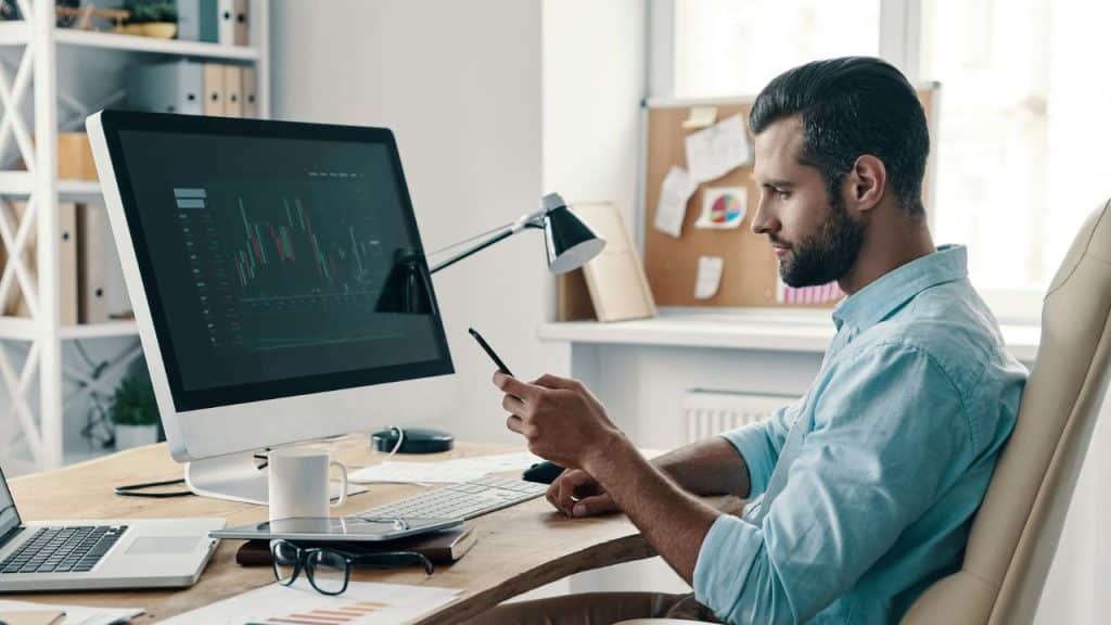 A man sits at a desk, looking at his phone while monitoring a large computer screen.