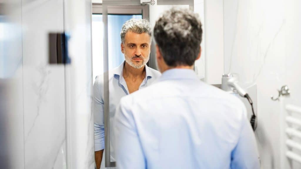 A man with a salt-and-pepper beard looks intently at his reflection in a mirror.