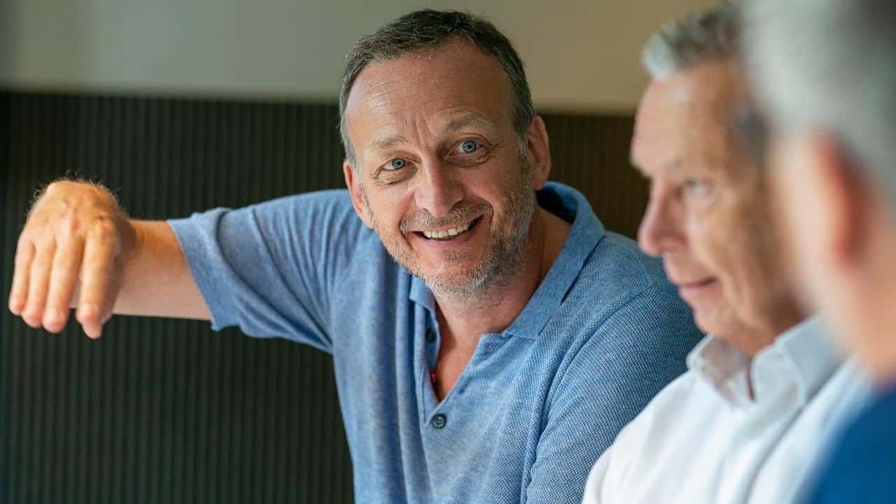 A smiling man in a blue shirt talks with others.