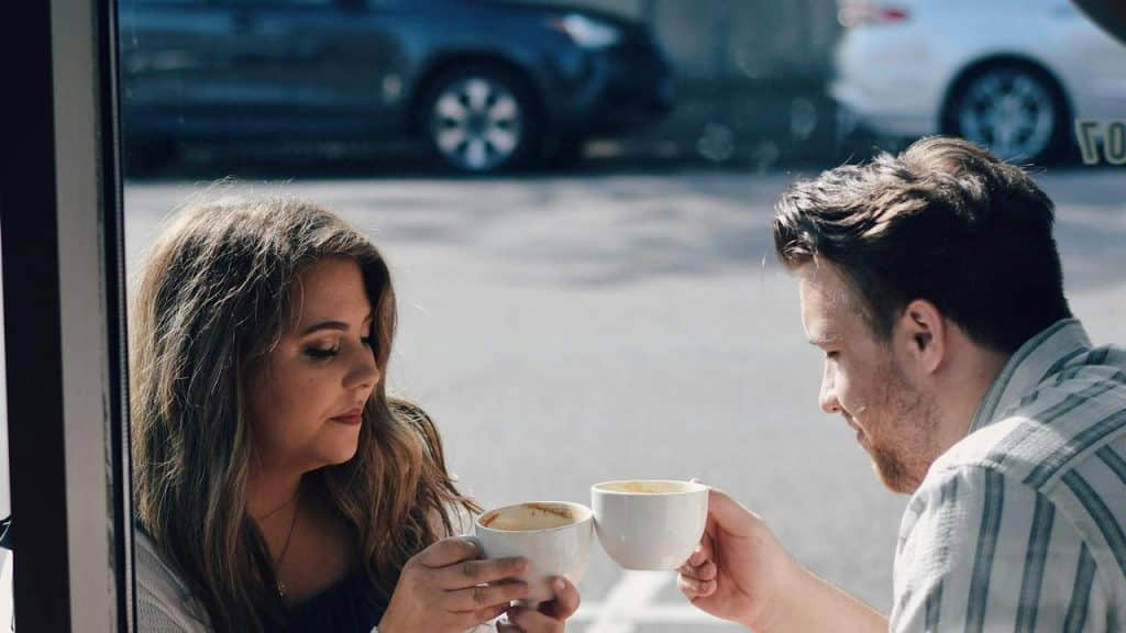 A couple clinking coffee mugs at a café table.