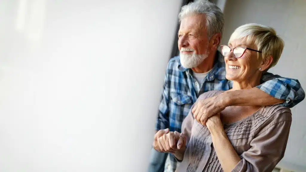 A happy senior couple looks out a window, with the man's arm around the woman.