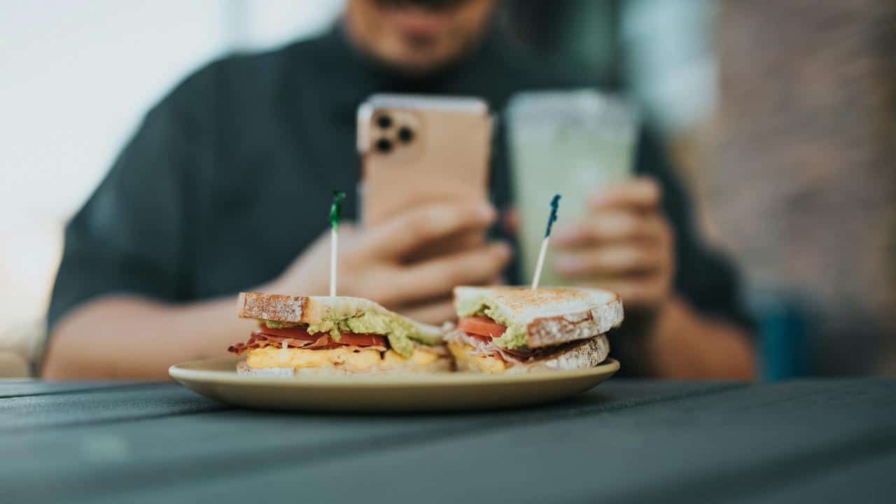 A sandwich on a plate with a man holding a phone and drink.