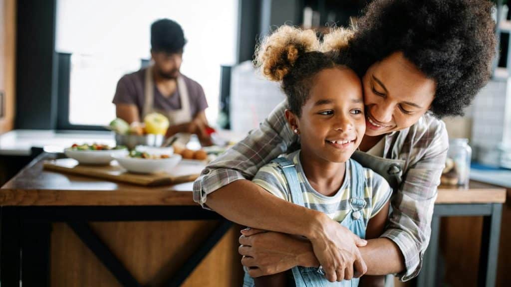 A mother hugging her daughter in the kitchen while someone cooks in the background.