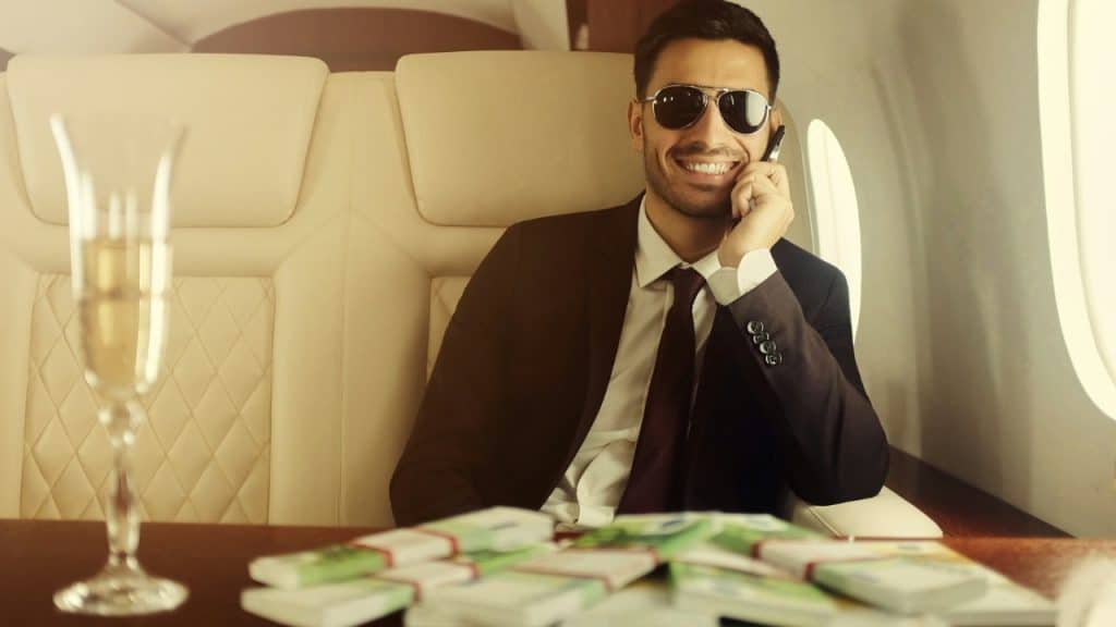 A smiling man in sunglasses and a suit talks on a phone in a private jet, with money on the table.