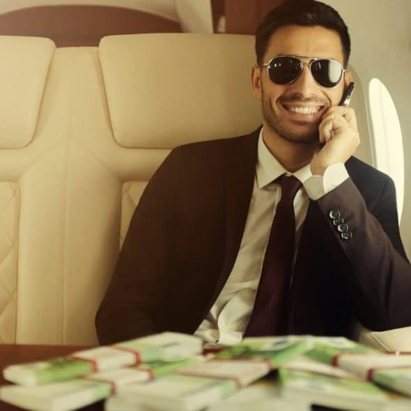 A smiling man in sunglasses and a suit talks on a phone in a private jet, with money on the table.