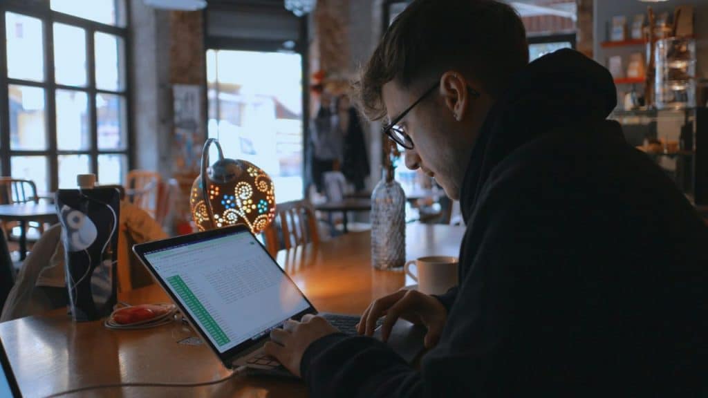 A person wearing glasses and a dark hoodie works on a laptop at a cafe table.