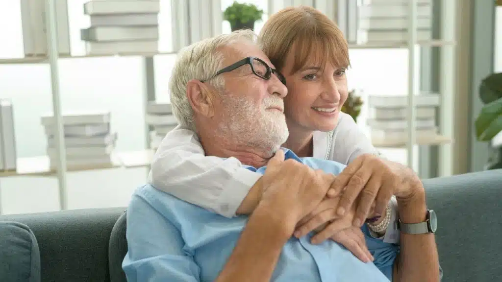 A senior woman with auburn hair hugs a smiling senior man with a white beard from behind.