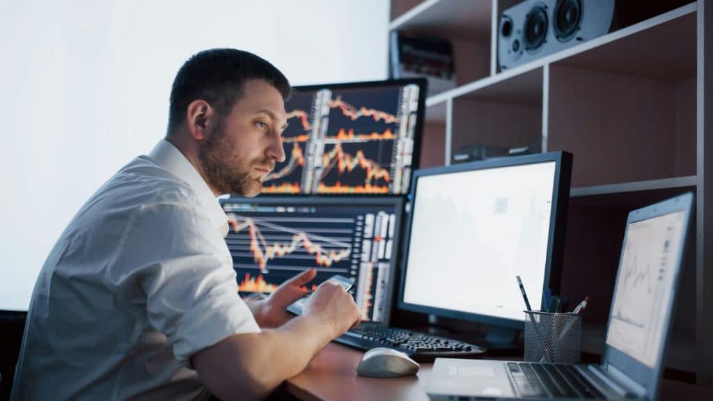 A man in a white shirt looks at multiple computer screens displaying financial charts.