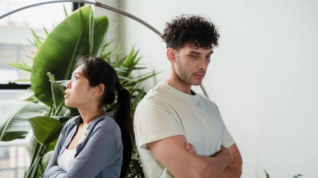 A displeased couple stands back-to-back indoors, arms crossed, avoiding eye contact.