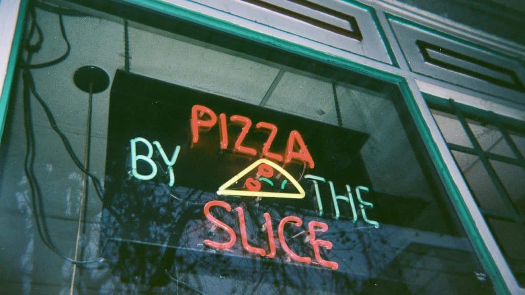 A neon sign reading "Pizza by the Slice" with a pizza slice graphic.