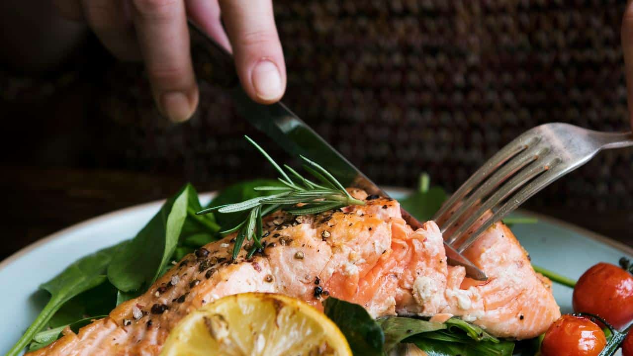 A person cutting into a grilled salmon fillet with greens and lemon.