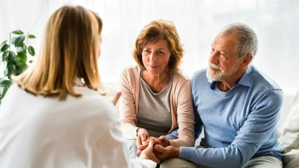A female healthcare professional consults with a senior couple, who are holding hands and listening attentively.