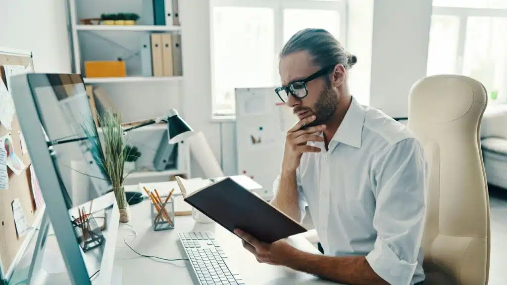 A focused man in glasses and a white shirt reads a book at his desk.