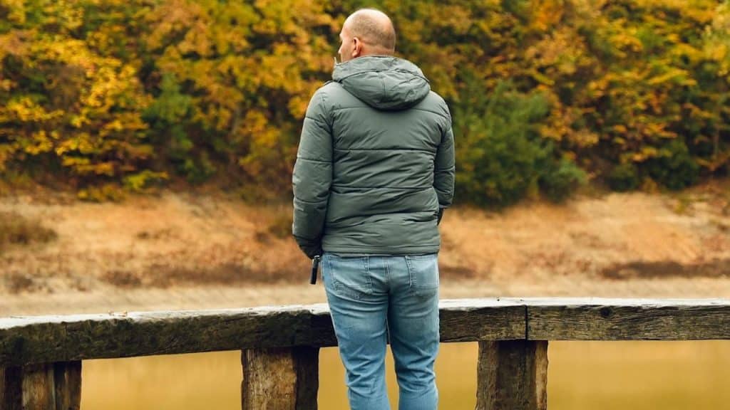 A man in a green puffer jacket and jeans stands, looking out over a lake with autumn trees.