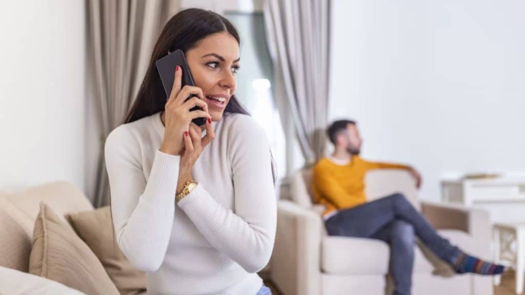 Woman smiling while on the phone with a friend, husband watching from the background