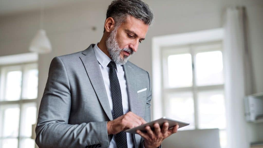 A professional man with a beard and grey suit focuses on a tablet in his hands.