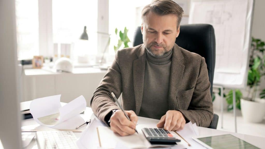 A bearded man in a brown jacket and turtleneck calculates on a desk.