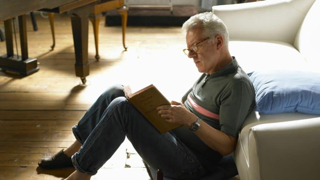 A mature man with glasses sits on the floor, engrossed in reading a book.