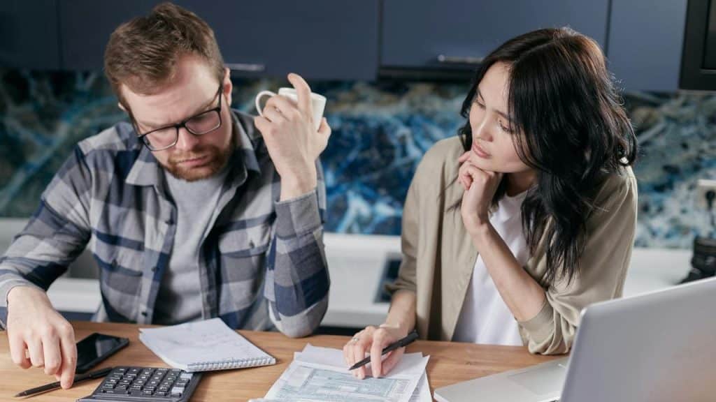 A serious couple reviews finances at a table with a laptop, calculator, and papers.