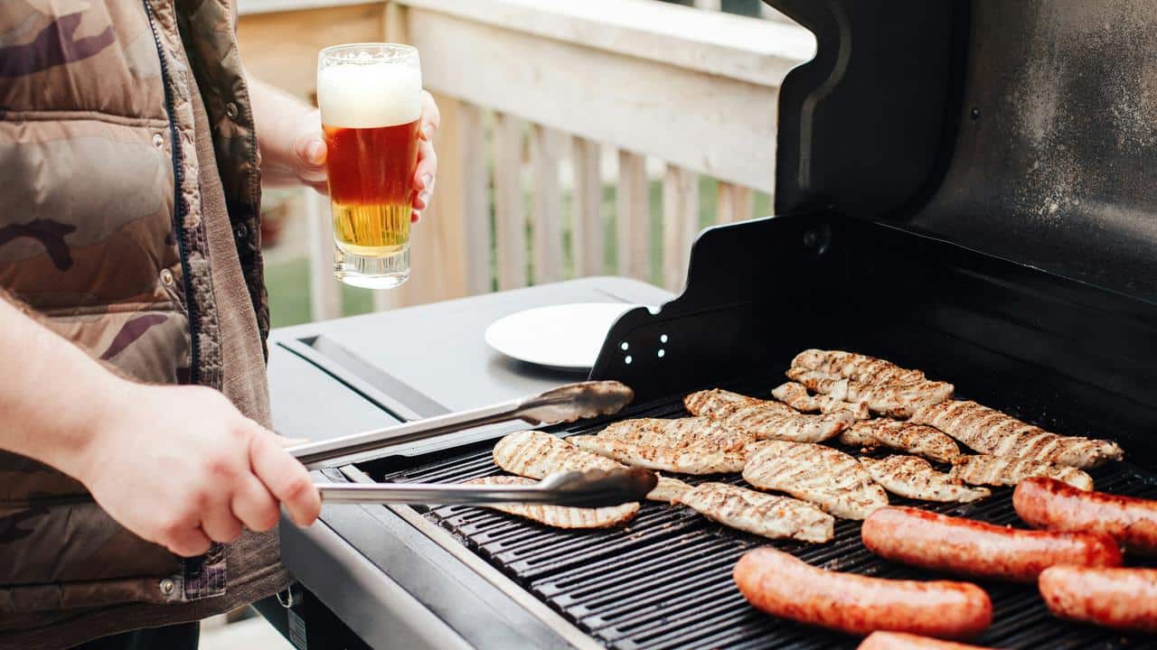 A man grills meat while holding a glass of beer.