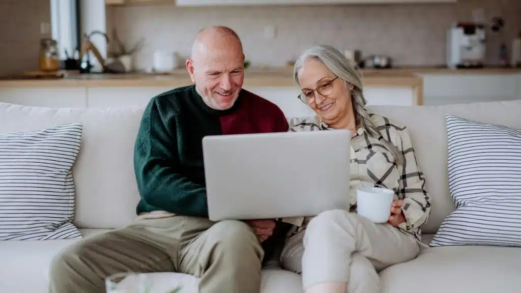 A smiling senior couple sits on a couch, looking at a laptop together, with a cup nearby.