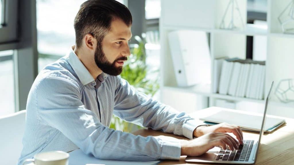 A focused man with a beard and striped shirt works on a laptop at a desk in a bright office.