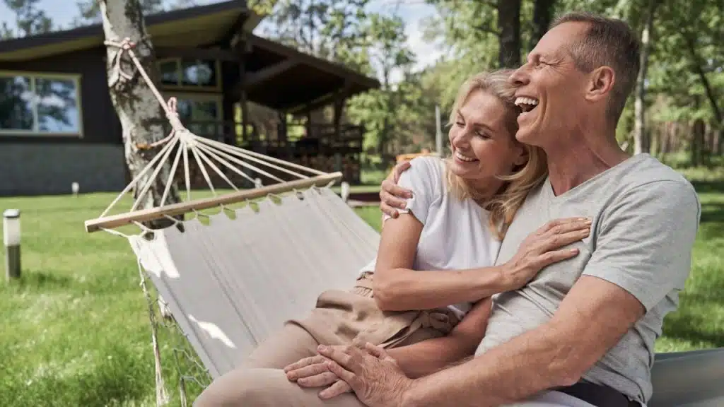 A laughing couple sits embraced in a hammock outdoors, with a house in the background.