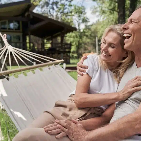 A laughing couple sits embraced in a hammock outdoors, with a house in the background.