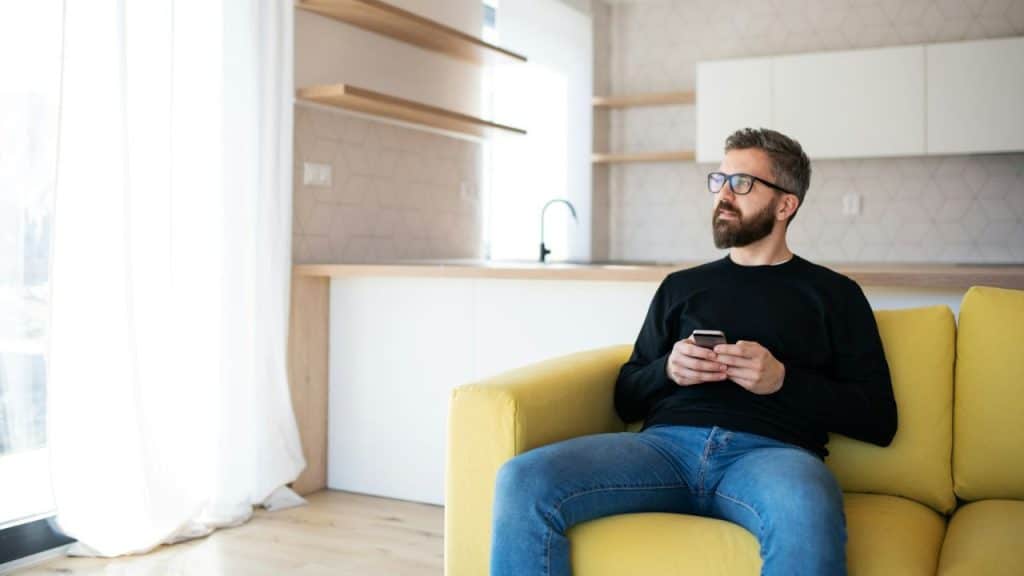 A bearded man in glasses and a black shirt sits on a yellow couch, looking away while holding a phone.